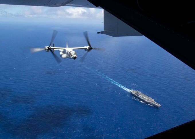A drone flies over the ocean, with a navy ship visible in the distance below.