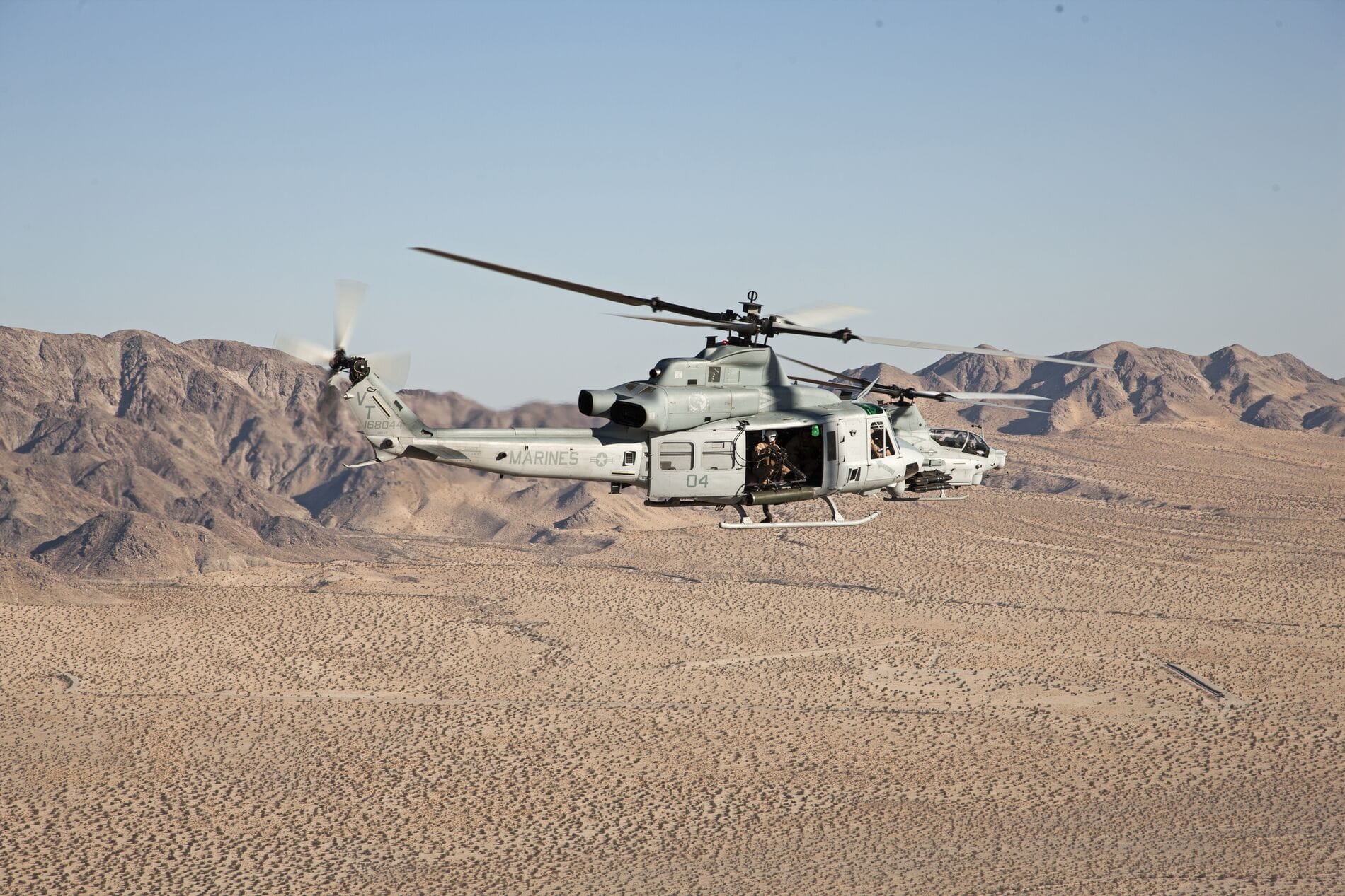 A military helicopter flies over a desert landscape with mountains in the background.