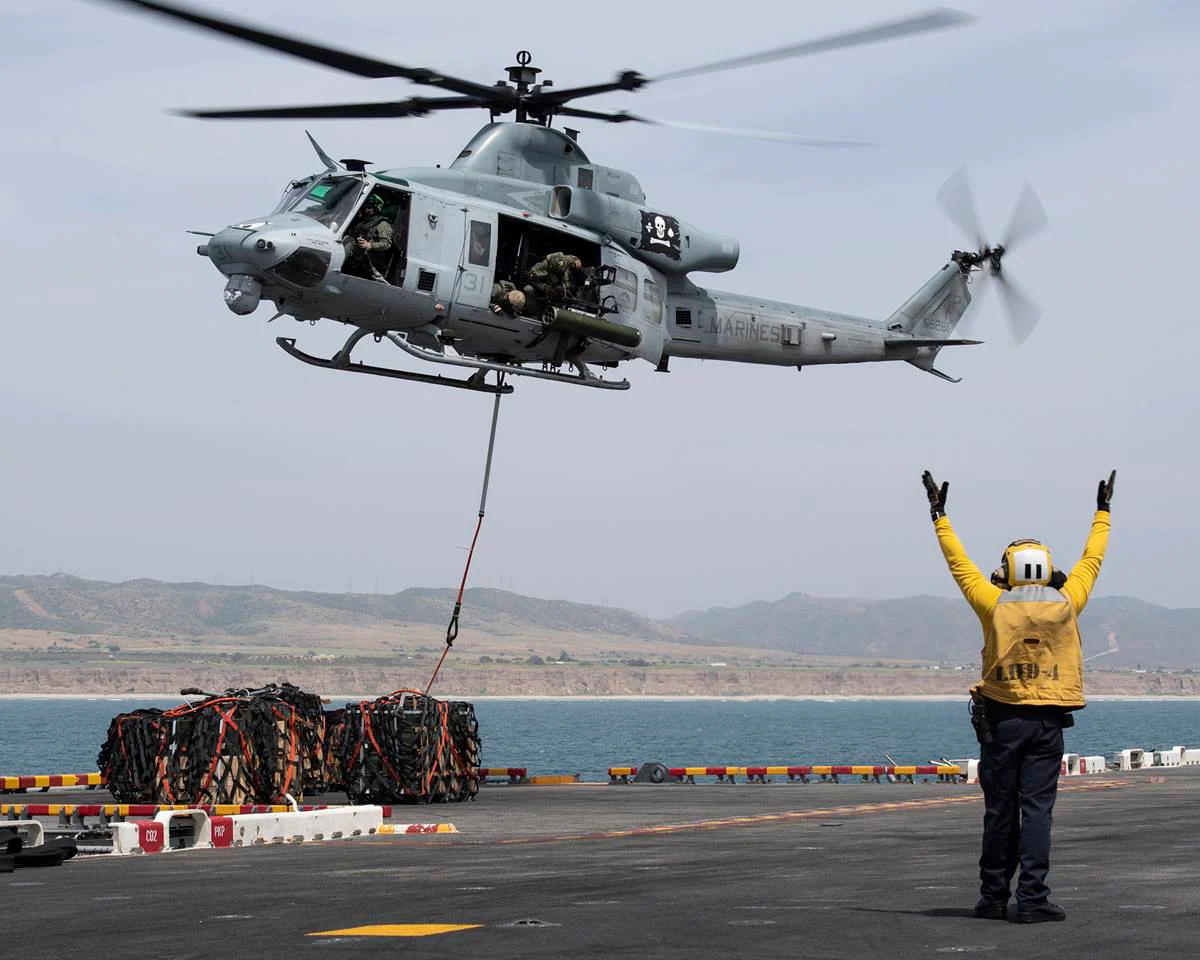 A Marine helicopter hovers above a ship while a crew member signals below. Cargo is suspended from