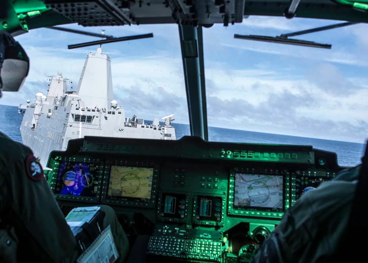 Cockpit view of a military helicopter flying close to a naval ship at sea with digital screens