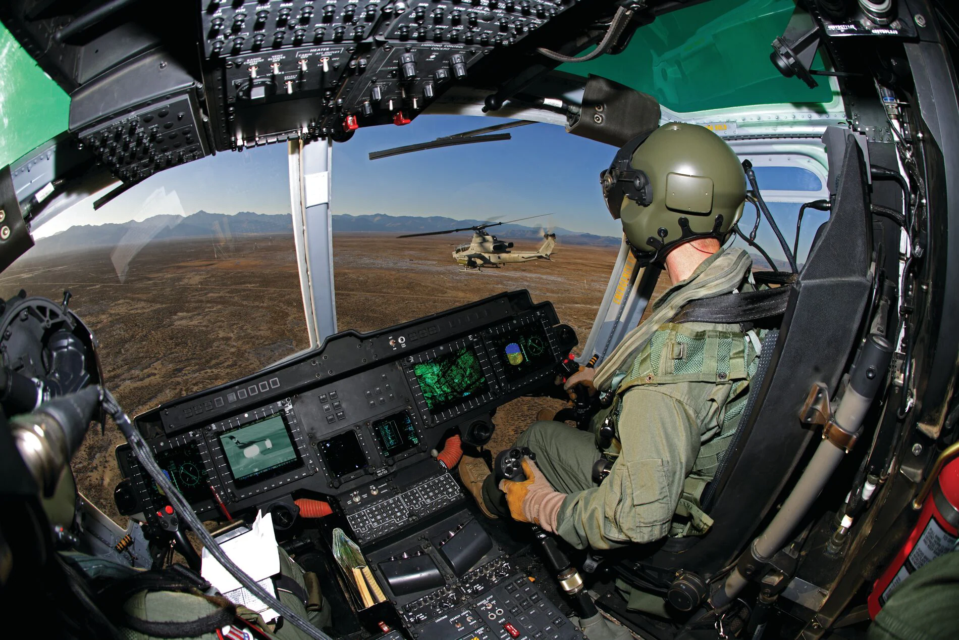 A pilot in a helicopter cockpit navigates over a mountainous landscape.