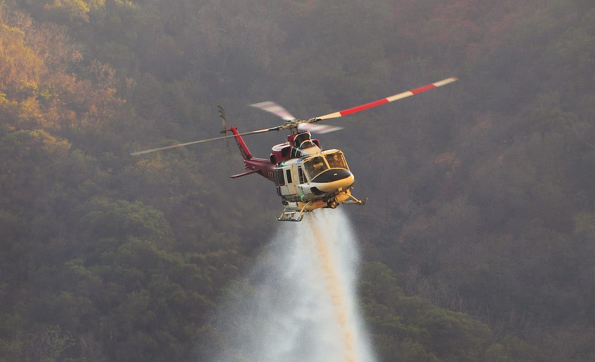 A helicopter spraying chemicals flies over a forested landscape. Smoke trails descend below.