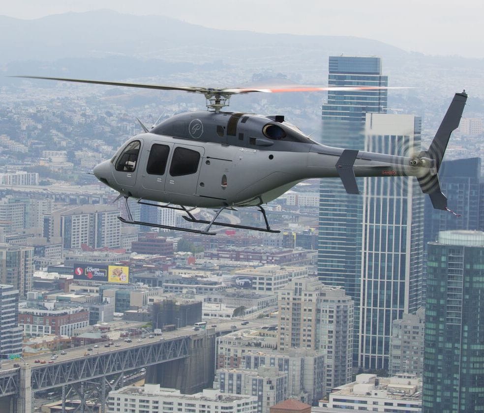 A gray helicopter flying over an urban skyline with skyscrapers and a bridge below.