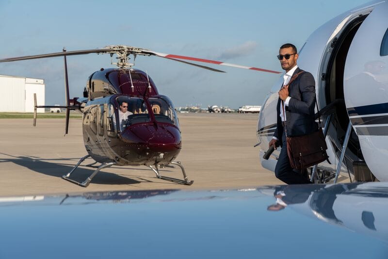 A man in a suit exits a private jet, with a helicopter parked nearby on the tarmac.