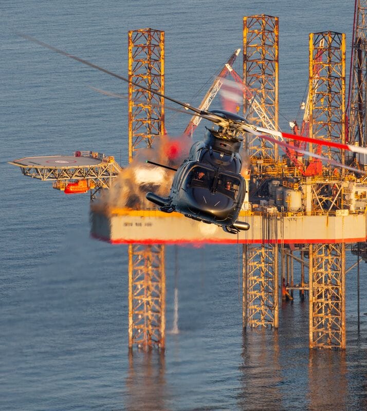 A helicopter flies near an offshore oil rig, with smoke and flames billowing from the structure.