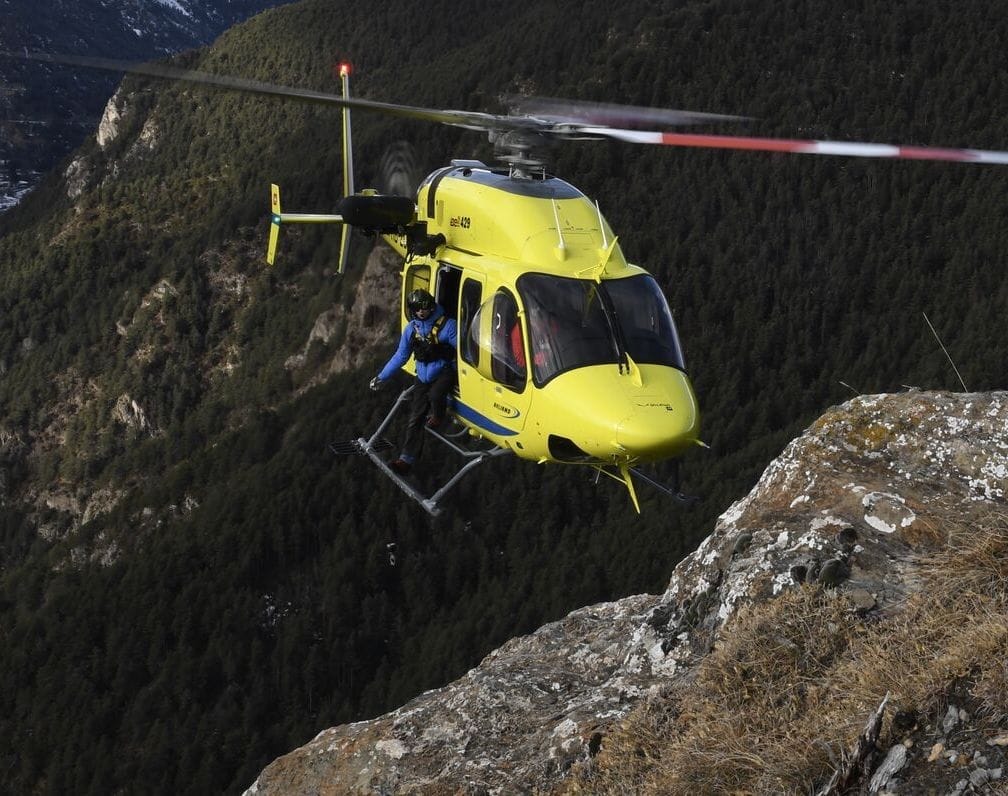 A yellow helicopter hovers near a steep rocky cliff with a person being lowered on a rescue line.