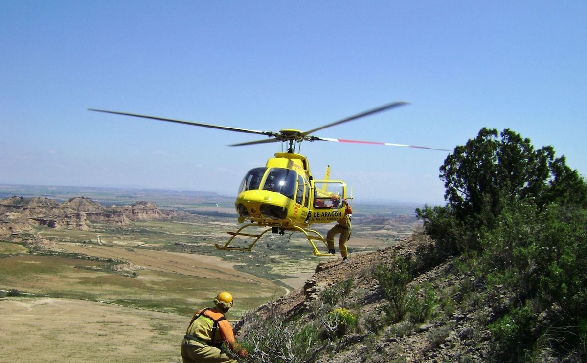 A yellow helicopter hovers near a rocky terrain, with a rescuer preparing to descend.