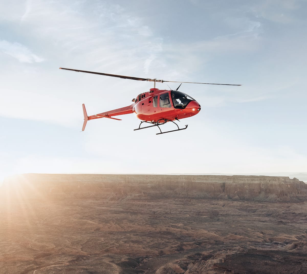 A red helicopter flying above a vast, rocky landscape under a clear sky.