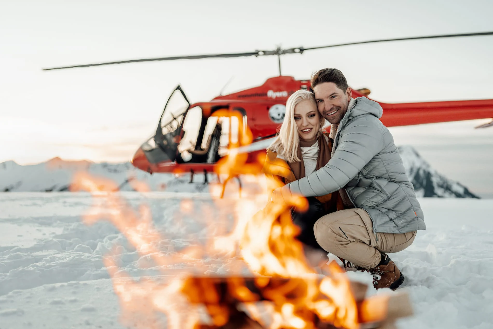 Couple sitting by a fire in the snow, with a helicopter in the background at sunset.