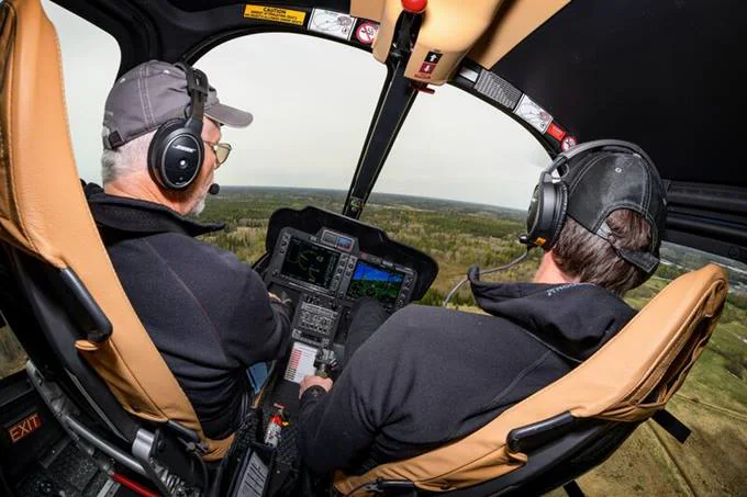 Two pilots in a helicopter cockpit, focused on the controls and the landscape below.