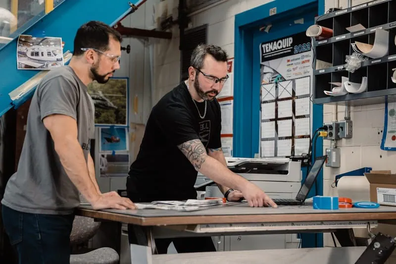 Two men are working together at a desk, discussing items displayed on a laptop screen.