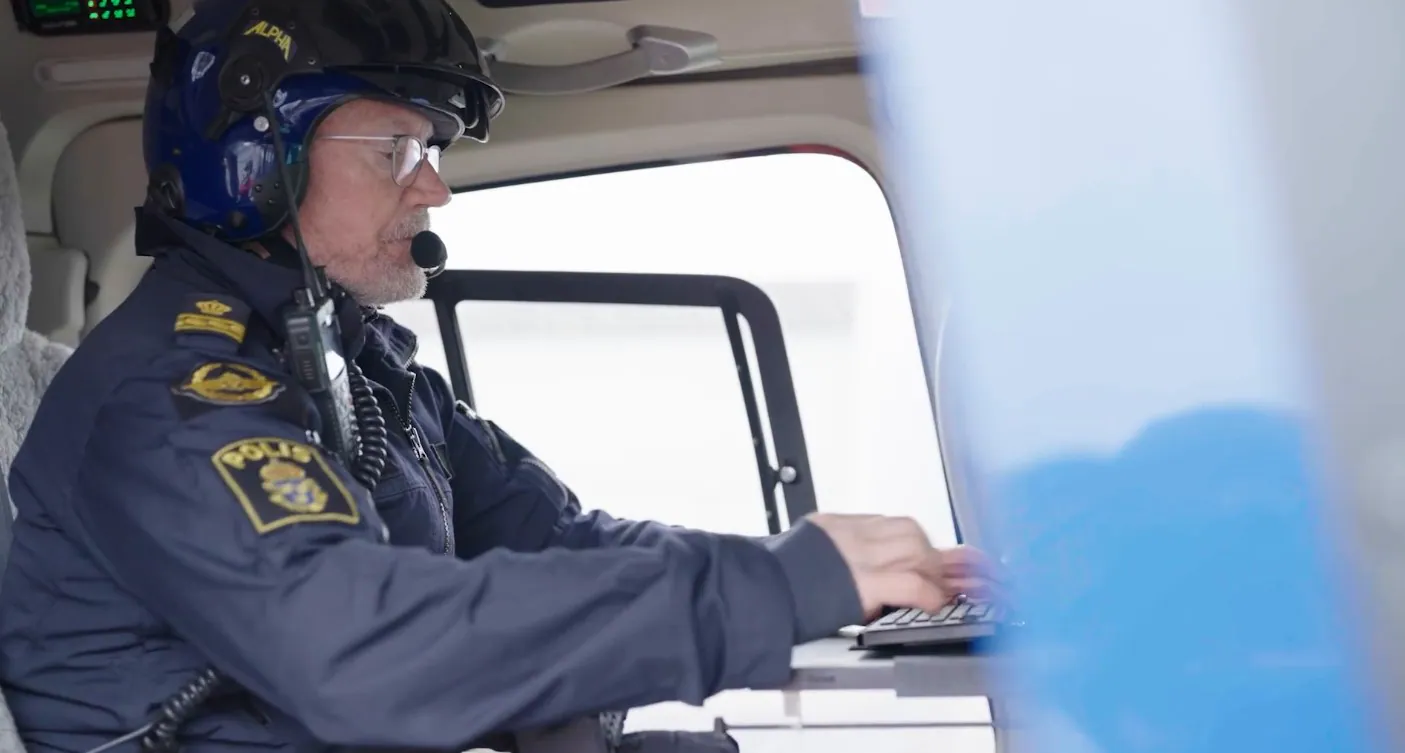 A man in a helmet and uniform typing on a laptop inside a helicopter cockpit.