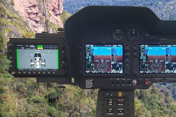 Cockpit display of a helicopter with multiple screens showing flight data and terrain in background.