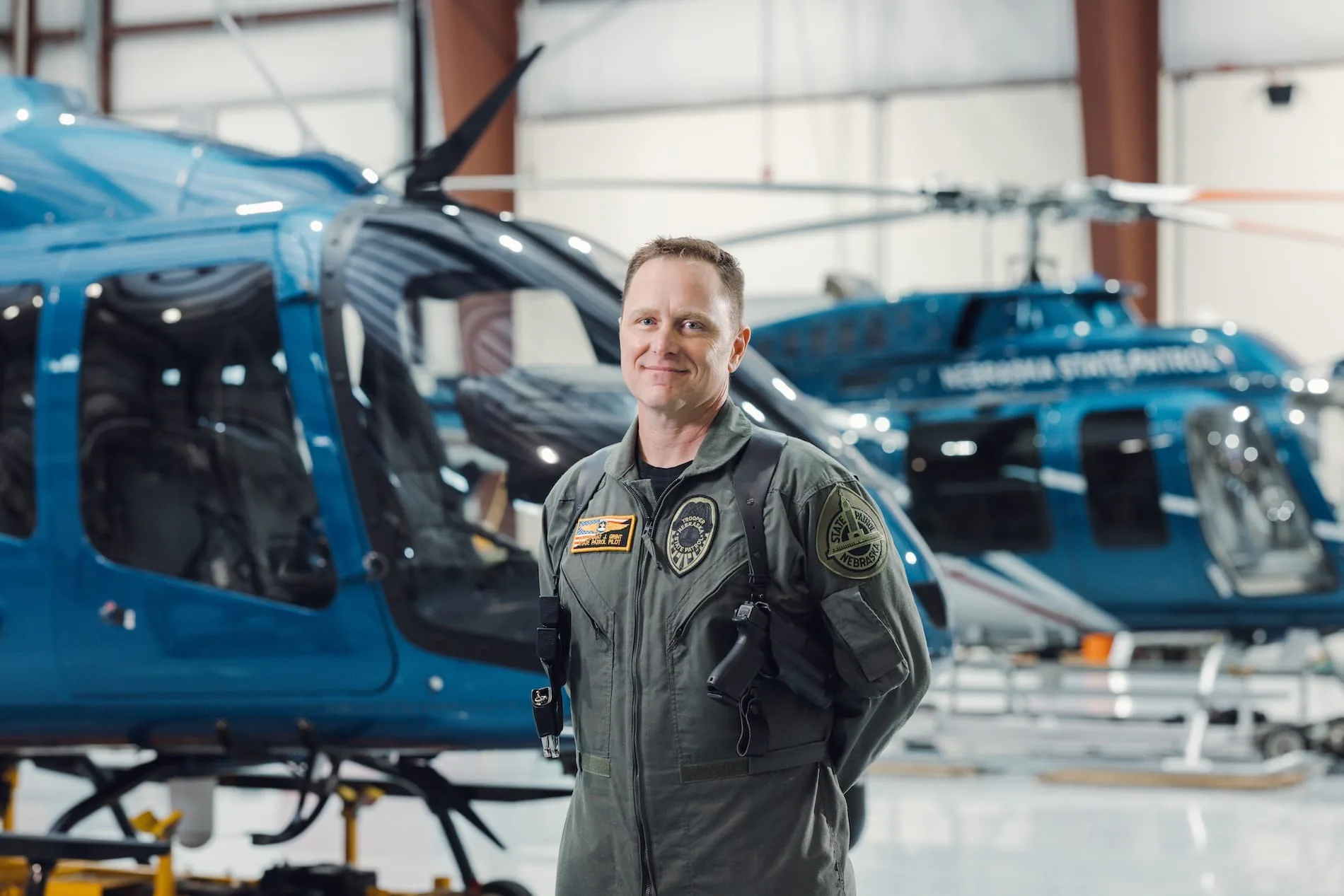 A smiling helicopter pilot in a flight suit stands between two blue helicopters in a hangar.