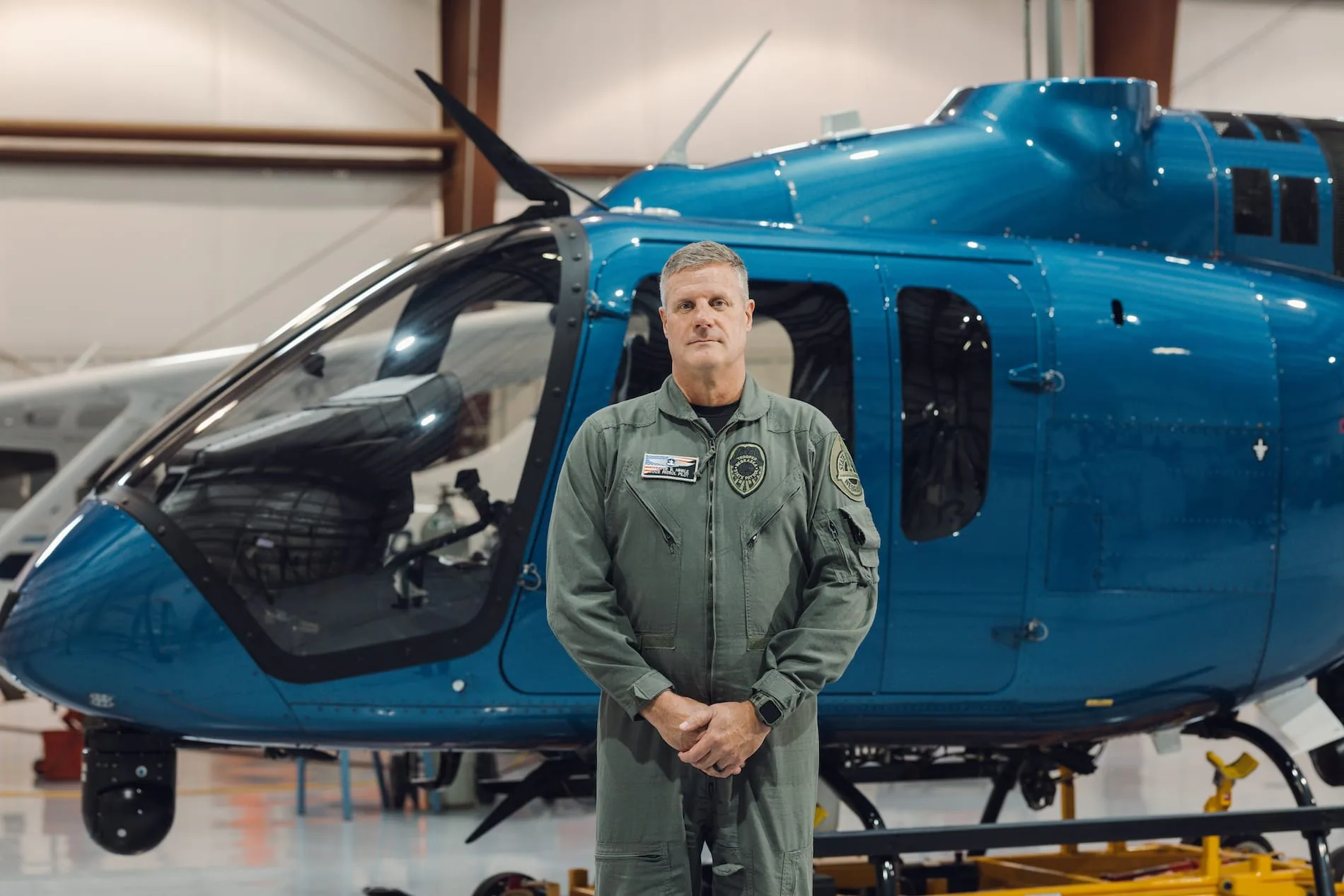 A male pilot in a green flight suit stands next to a blue helicopter in a hangar.