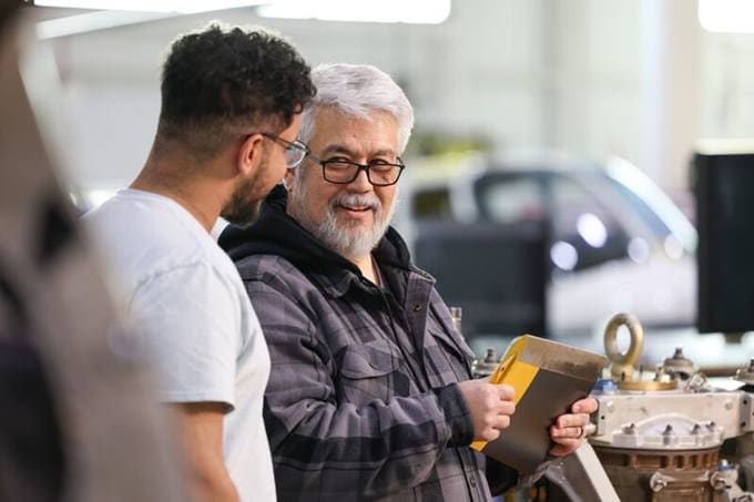 Two men in a workshop discuss a metal part; one holds a yellow object, both are smiling.