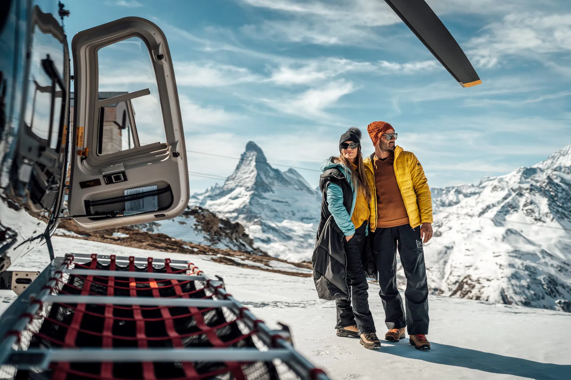 Two people stand on snow-covered mountainside beside a helicopter, with peaks in the background.