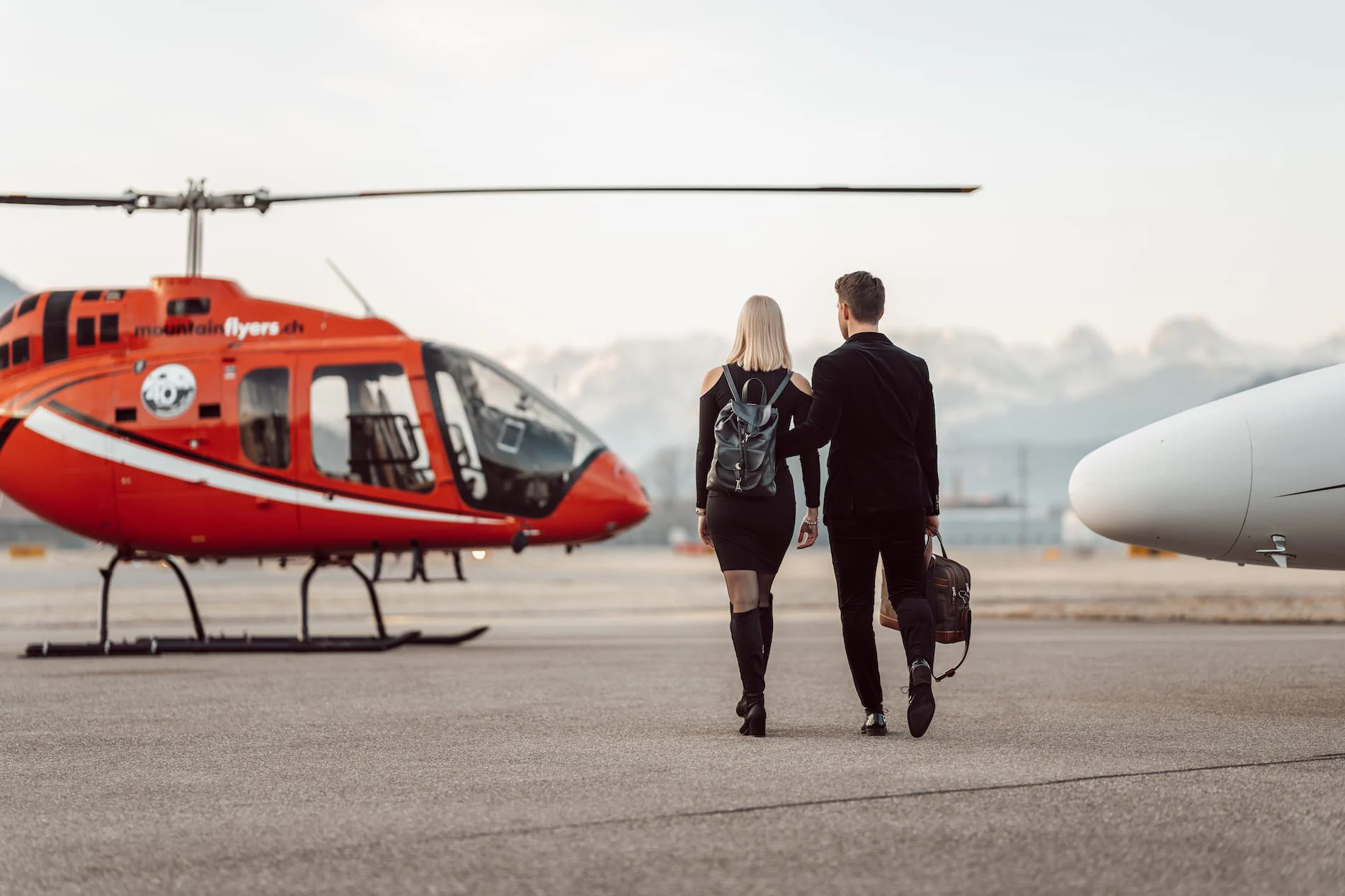A couple walks towards a red helicopter at an airfield, with mountains in the background.