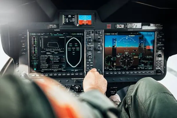 Cockpit controls with screens displaying flight data and a pilot's hand on the control panel.