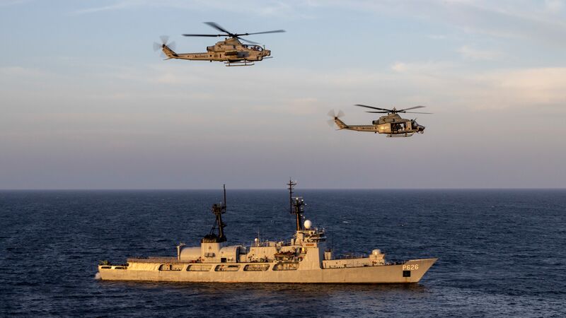 Two helicopters fly over a naval vessel in the ocean during sunset.