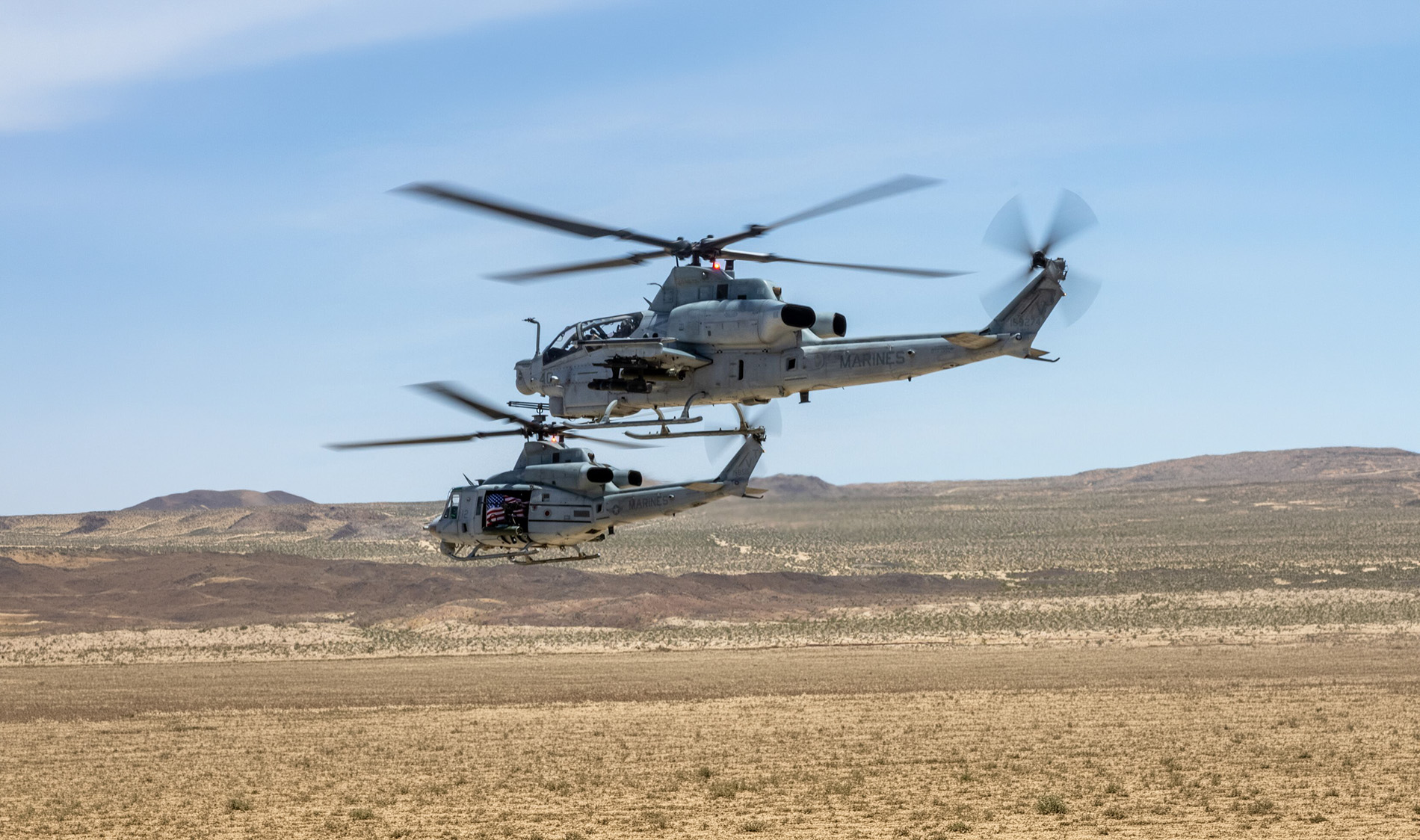 Two military helicopters flying in a desert landscape under a clear blue sky.