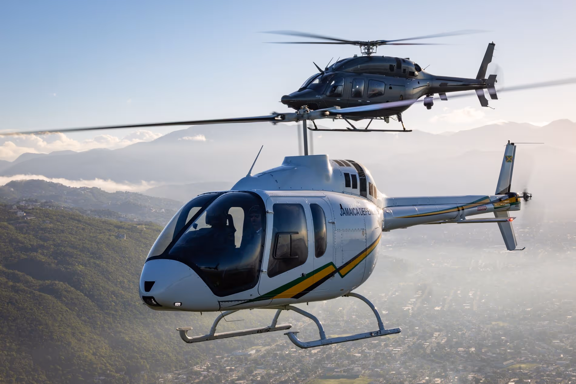 Two Jamaica Defence Force helicopters flying over mountainous terrain and cityscape in daylight.