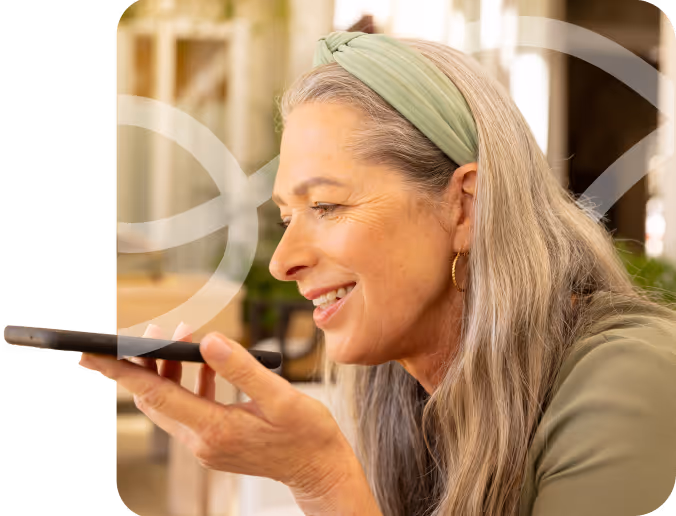 Smiling woman with gray hair and a green headband speaking into a smartphone held close to her mouth.