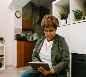 An elderly man wearing glasses and a plaid shirt works on a laptop at a desk, with an older woman standing behind him in a bright, modern living room.