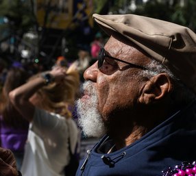 Older man with sunglasses and a hat standing outside in a crowd of people on a sunny day