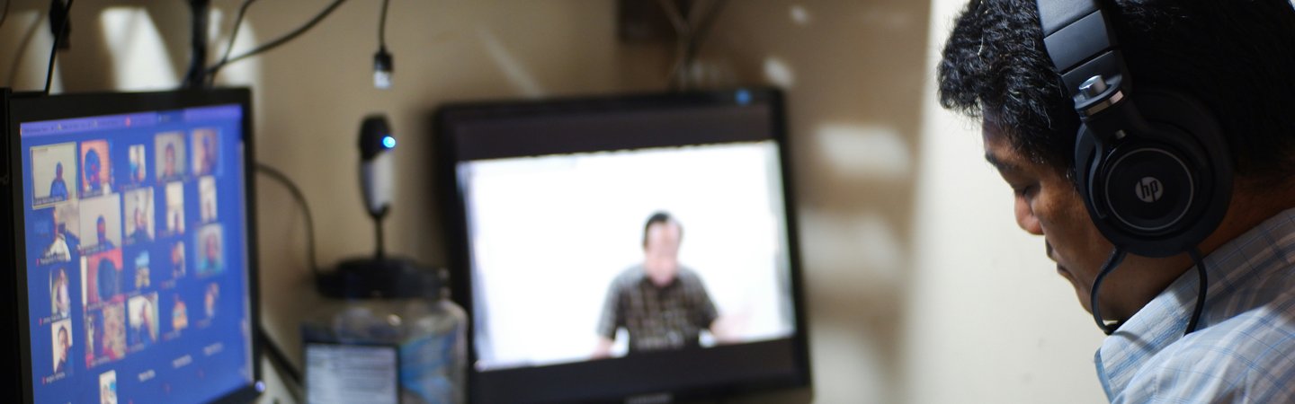A man wearing headphones takes notes during an online meeting with two computer monitors at a cluttered desk.