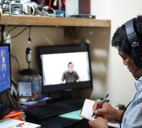 A man wearing headphones takes notes during an online meeting with two computer monitors at a cluttered desk.