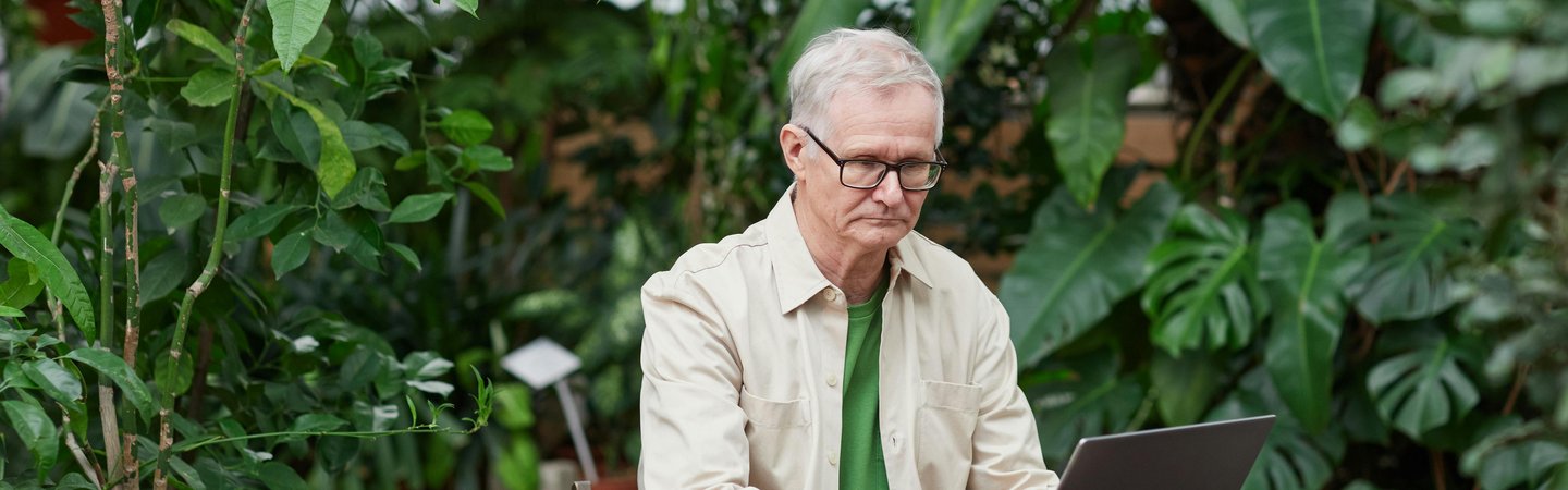 Older man working on his laptop at a desk surrounded by trees.
