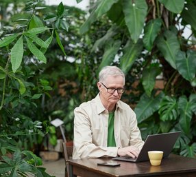 Older man working on his laptop at a desk surrounded by trees.