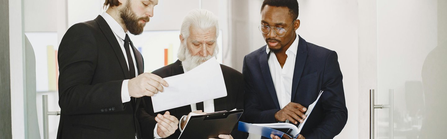 Three professional men in suits stand together in an office hallway, reviewing documents and a clipboard in a collaborative discussion.