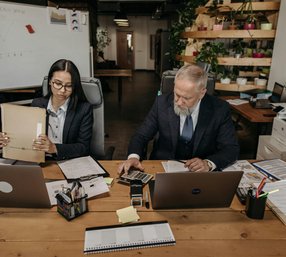 A younger woman and a senior male colleague in suits sit side by side at a busy office desk, reviewing documents and working on laptops with a quarterly report whiteboard in the background.