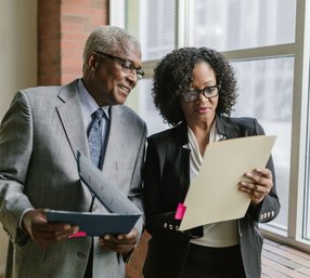 A senior businessman and a professional woman in suits stand together near a window, reviewing documents from folders in an office hallway.