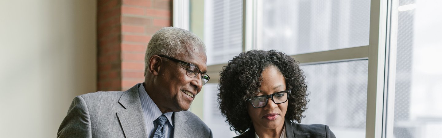 A senior businessman and a professional woman in suits stand together near a window, reviewing documents from folders in an office hallway.