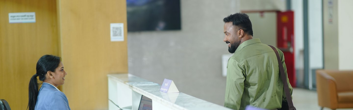 A receptionist in a blue blazer smiles while helping a visitor at a bright, modern front desk in an office lobby.