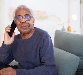 A senior man wearing glasses sits on a couch at home, holding a phone to his ear with a thoughtful expression.