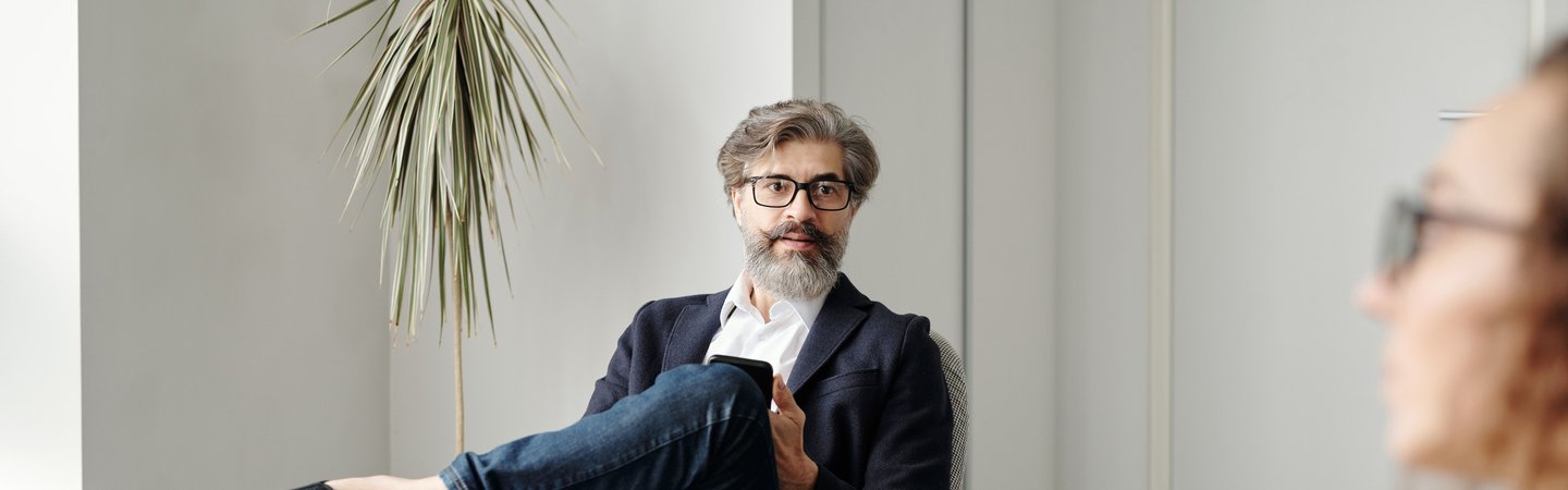 A bearded man in glasses and a blazer sits relaxed in a modern office chair during a conversation, with another person visible in the foreground holding a notepad.