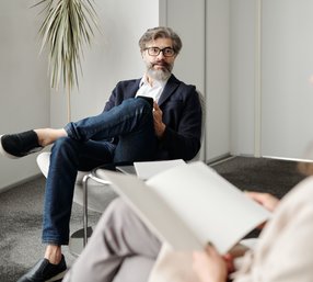 A bearded man in glasses and a blazer sits relaxed in a modern office chair during a conversation, with another person visible in the foreground holding a notepad.