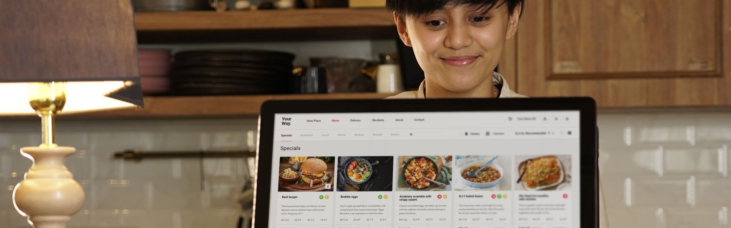 A smiling restaurant worker stands behind a modern touchscreen POS system displaying a digital menu with food specials in a cozy kitchen setting.