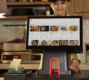 A smiling restaurant worker stands behind a modern touchscreen POS system displaying a digital menu with food specials in a cozy kitchen setting.