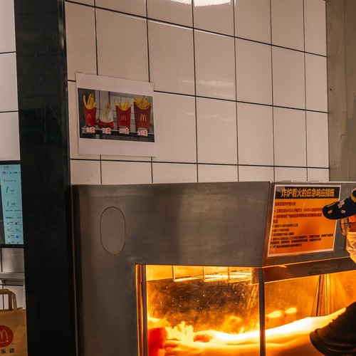A kitchen worker in a black uniform and face mask reaches into a stainless steel fry station under warm overhead lighting in a busy commercial kitchen.