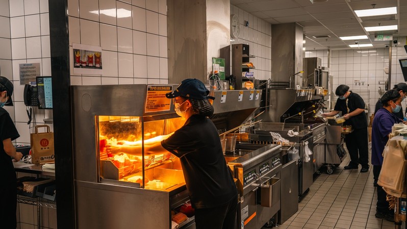 A kitchen worker in a black uniform and face mask reaches into a stainless steel fry station under warm overhead lighting in a busy commercial kitchen.