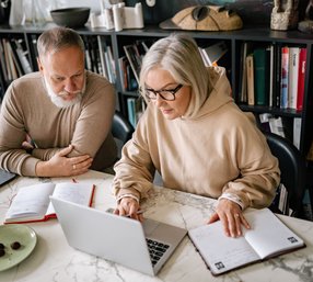 A mature man and woman sit together at a marble table, both focused on a laptop surrounded by open notebooks and a glass of juice in a cozy home office setting.