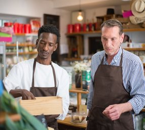 Two male retail workers in brown aprons stand together in a well-stocked shop, examining items from a wooden crate during a shift.
