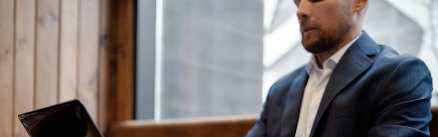 A man in a suit jacket types on a laptop at a wooden table in a warm, casual cafe setting.