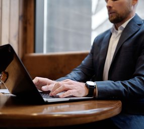 A man in a suit jacket types on a laptop at a wooden table in a warm, casual cafe setting.