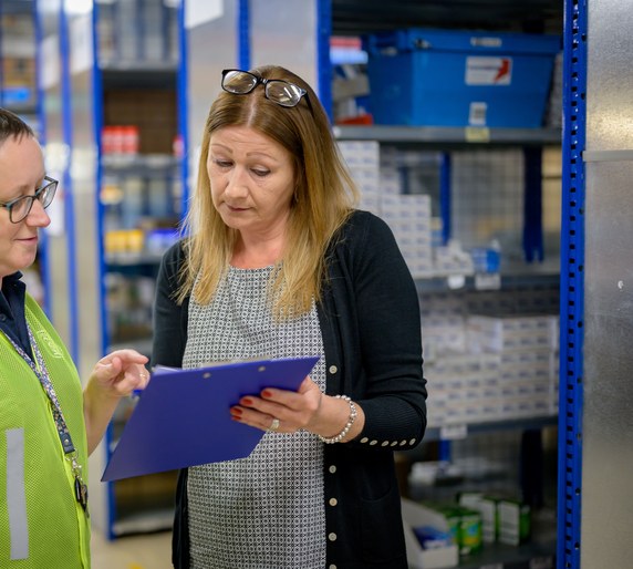 A warehouse team member in a hi-vis vest reviewing a clipboard with a colleague